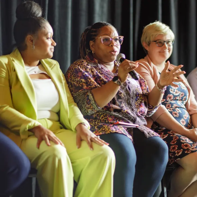 A woman speaking onstage, sitting next to two other women